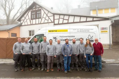Eine Handwerkergruppe steht vor einem Firmenwagen und einem Fachwerkhaus bei der Holzwerkstatt.